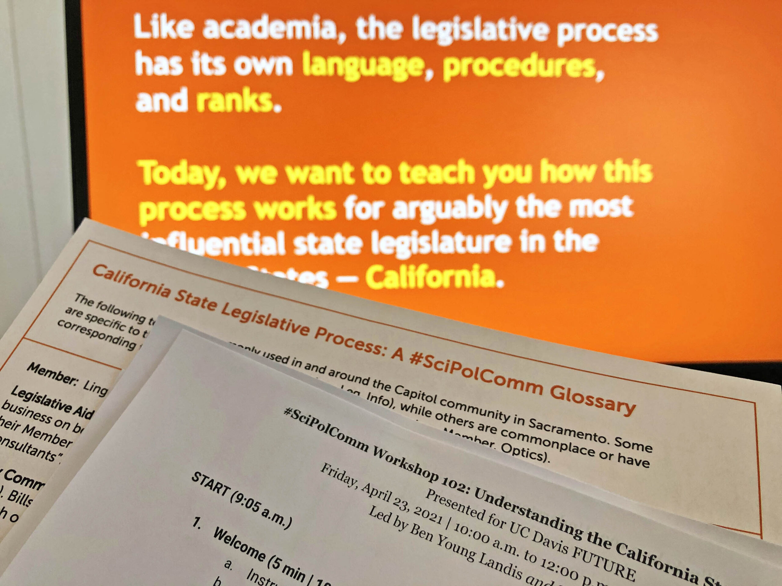 Photo of a pair of worksheets held up in front of a computer monitor. One worksheet reads: California State Legislative Process A hashtag Sci Pol Comm Glossary, while the other reads hashtag Sci Pol Comm Workshop 102. The monitor text is out of focus but reads "Like academia, the legislative process has its own language, procedures, and ranks. Today, we want to teach you how this process works for arguably the most influential state legislature in the United States em dash California