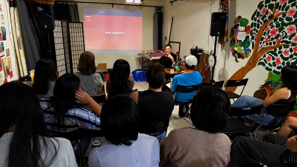 Photo of River Basham speaking to an audience smiling in front of a slide that reads Why a Sphere?